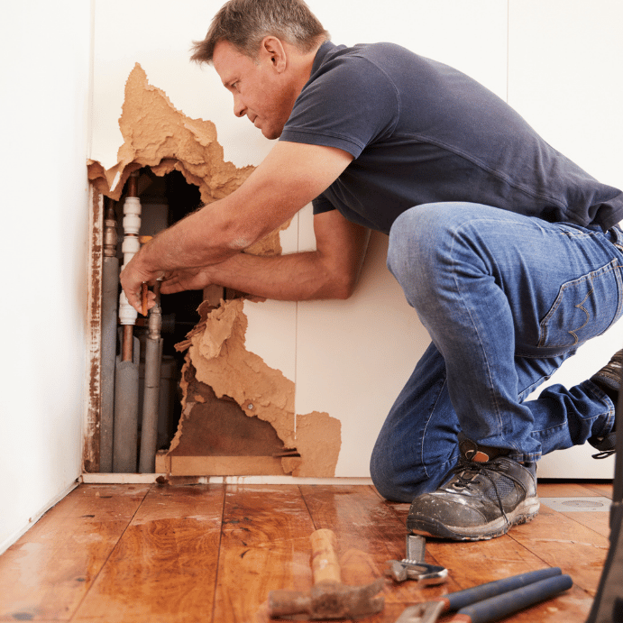Handyman kneels by a damaged wall, repairing exposed plumbing pipes behind torn drywall.