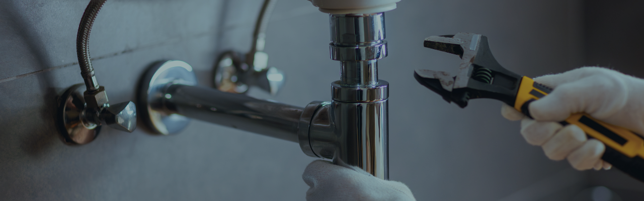 Gloved hands use an adjustable wrench to tighten a chrome drain trap under a sink during plumbing work