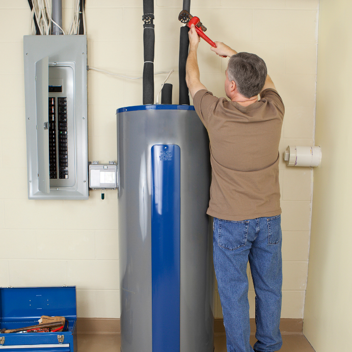 Maintenance worker tightening a valve on a tall water heater in a utility room by a toolbox and electrical panel.