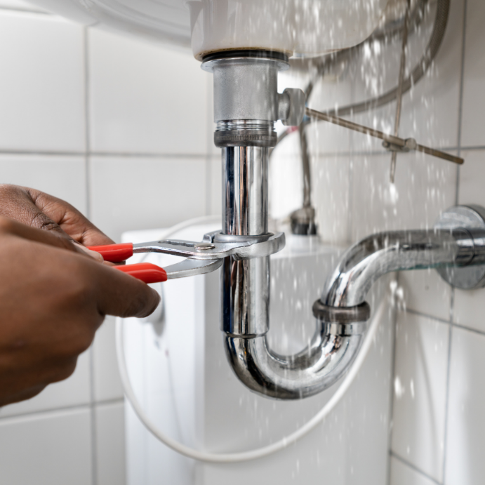 Close-up of hands using red-handled pliers to tighten a pipe under a sink with water spraying from a joint.