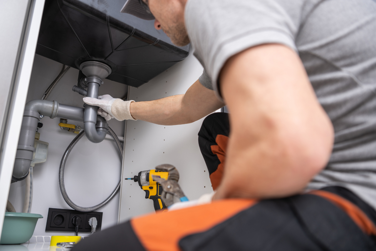 Plumber crouched under a sink, tightening a P-trap with gloved hand; cordless drill sits nearby on the cabinet shelf.
