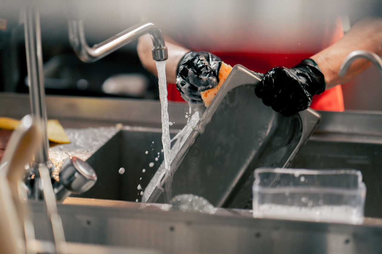 Gloved hands rinse a tablet under a steady stream of water in a kitchen sink.
