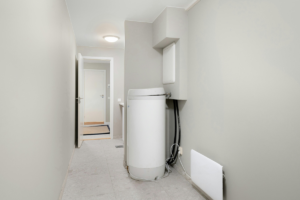 Narrow utility room featuring a tall white water heater, exposed hoses, and a wall cabinet. The hallway leads to a door at the far end.