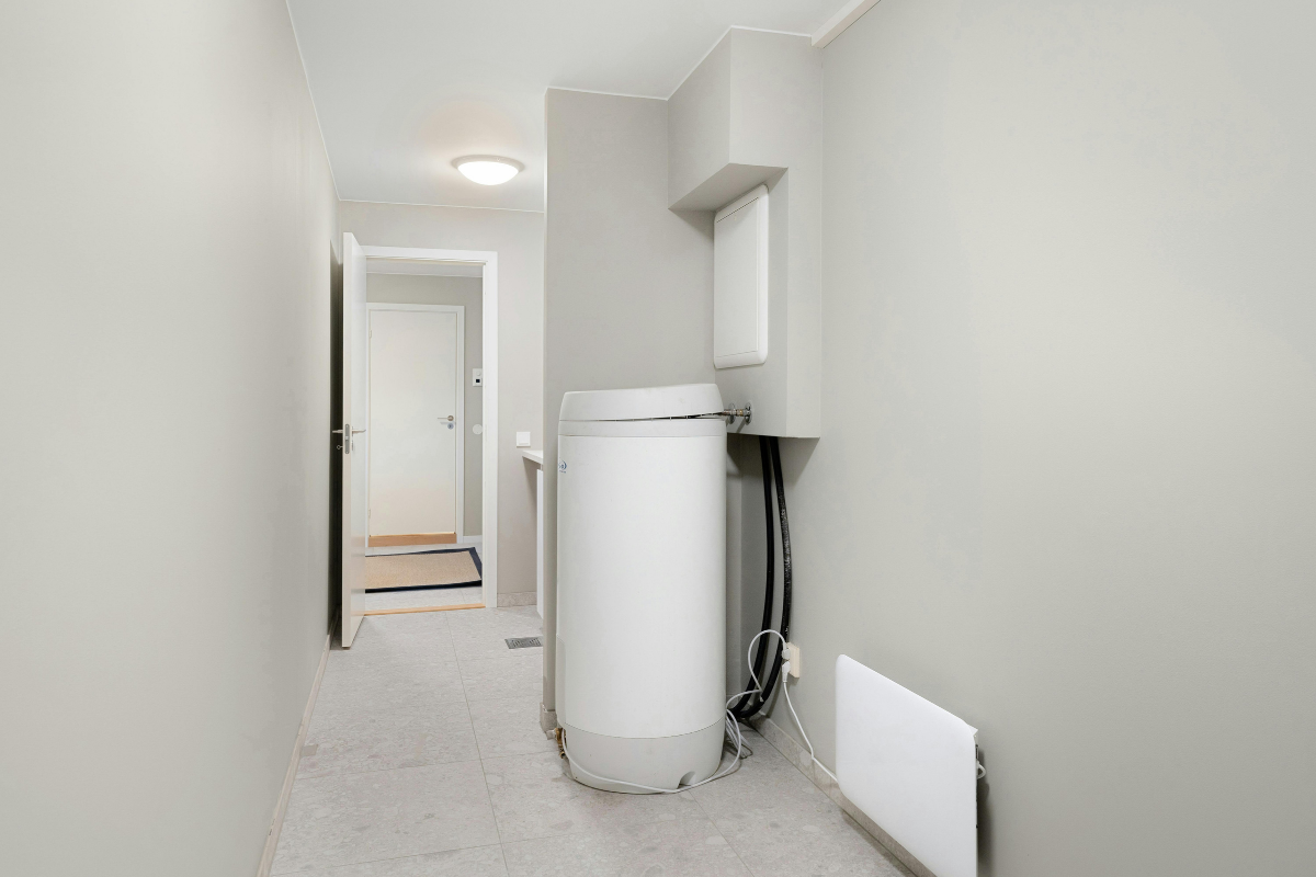 Narrow utility room featuring a tall white water heater, exposed hoses, and a wall cabinet. The hallway leads to a door at the far end.