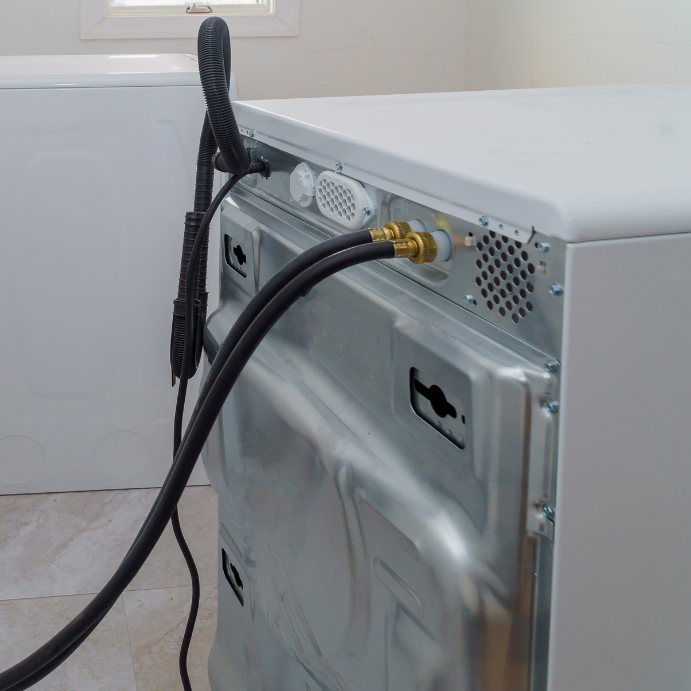 Back view of a washing machine with water supply hoses connected to brass fittings and a flexible drain hose spilling over the side of the unit with a bare room in the background.