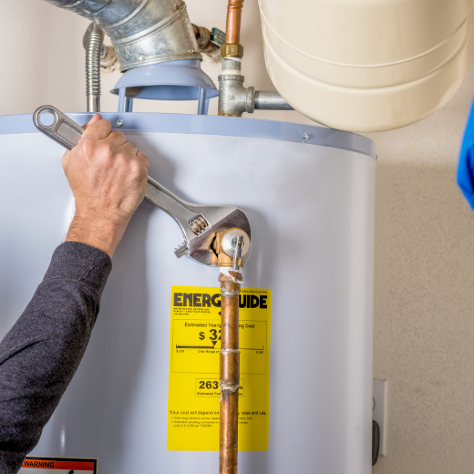 Person uses a wrench to tighten a valve on a vertical copper pipe connected to a water heater with an energy label visible in the foreground.