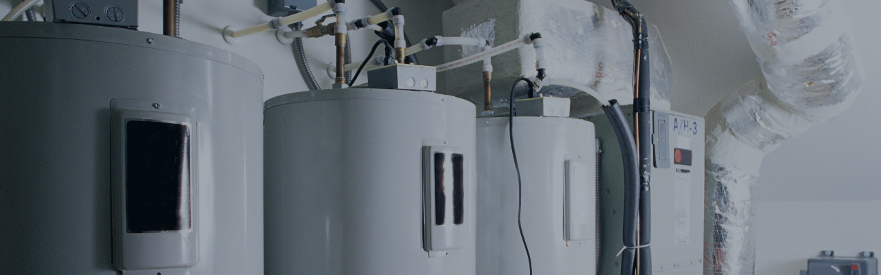 Row of large cylindrical industrial tanks with pipes and ducts in a mechanical room, likely heating equipment.