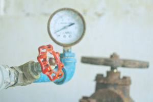 Industrial valve with a red wheel and pressure gauge in the background, blue piping shows wear and rust nearby.