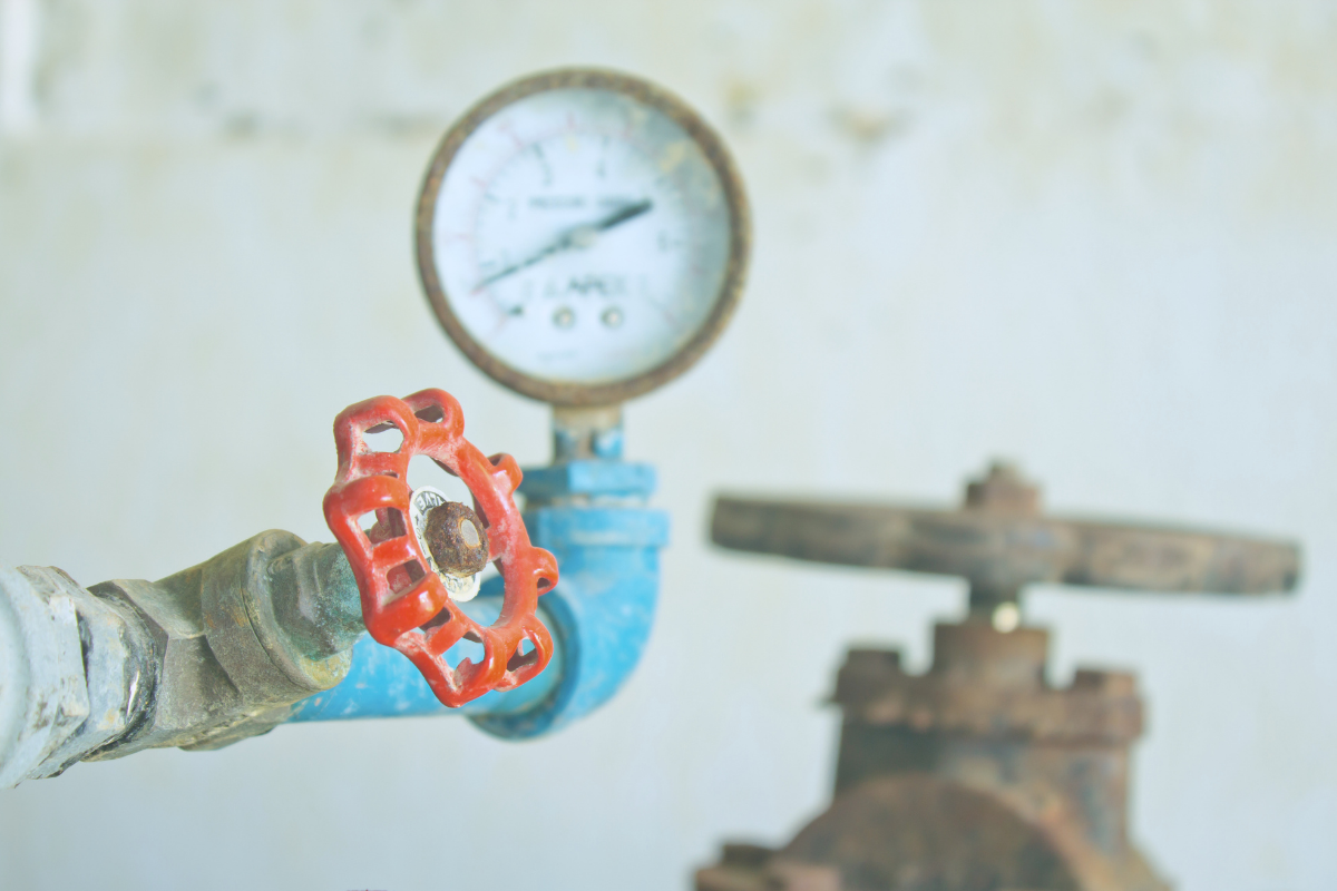 Industrial valve with a red wheel and pressure gauge in the background, blue piping shows wear and rust nearby.