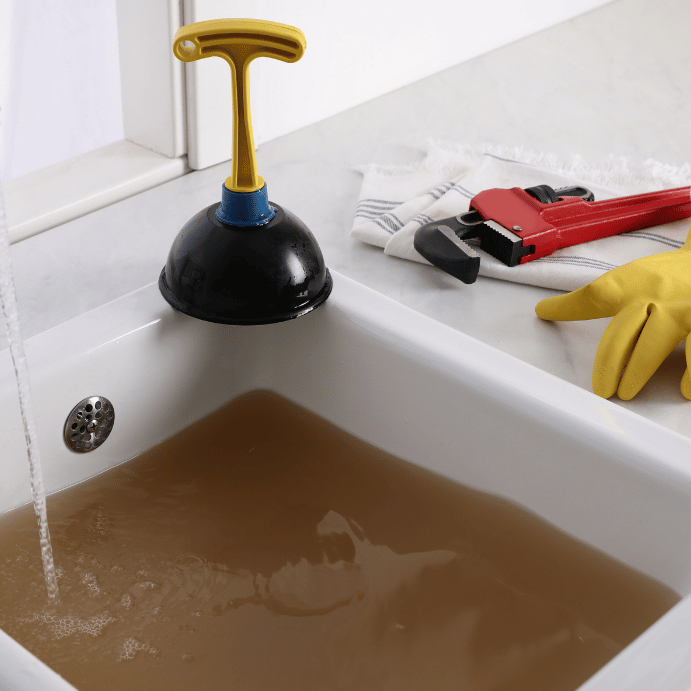 Plunger atop a bathtub edge with a brown-filled sink and repair tools nearby, ready for plumbing work (yellow gloves and red wrench).