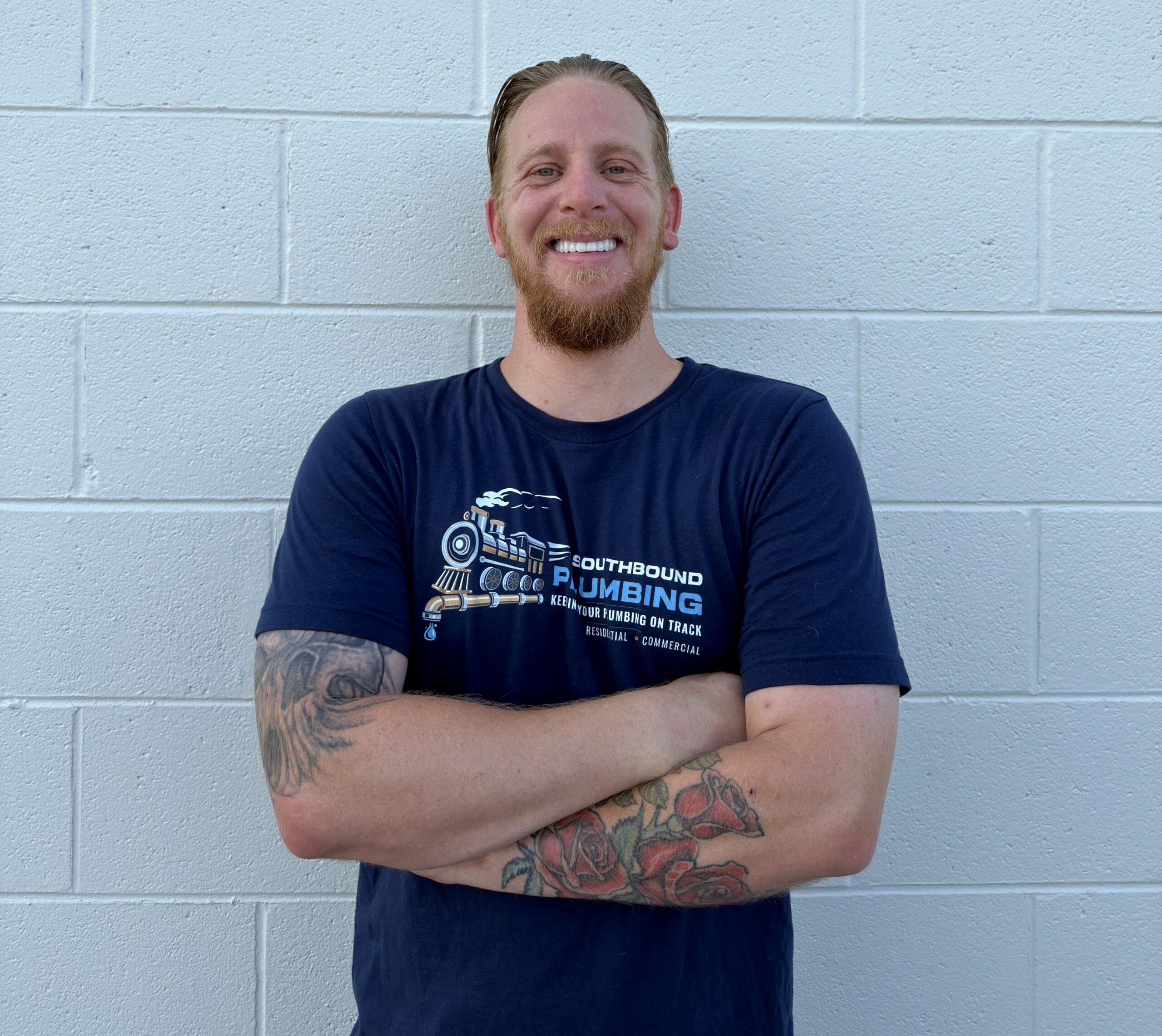 Man with a beard and tattoos standing with arms crossed, smiling against a light blue brick wall.