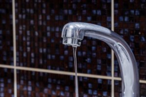 Close-up of a chrome faucet dripping a thin stream of water against a tiled wall background.