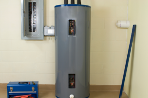 Tall gray vertical water heater in a utility room with panels open showing wiring, beside a blue toolbox and a wall-mounted panel on the left.