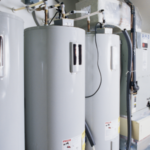 Row of gray industrial water heaters/boilers in a mechanical room with exposed pipes and wiring.